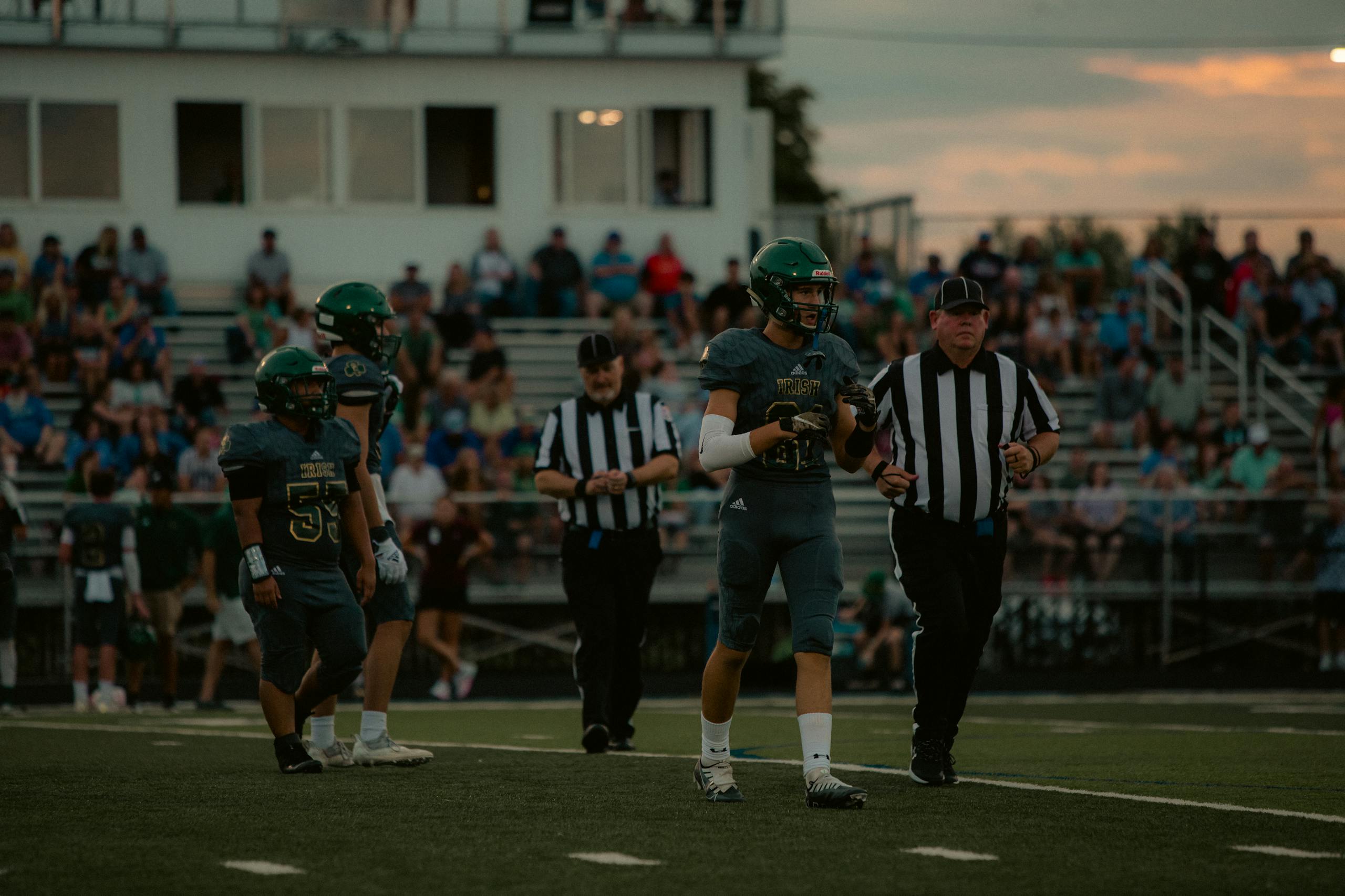 Players and referees on a football field during a high school game at sunset.